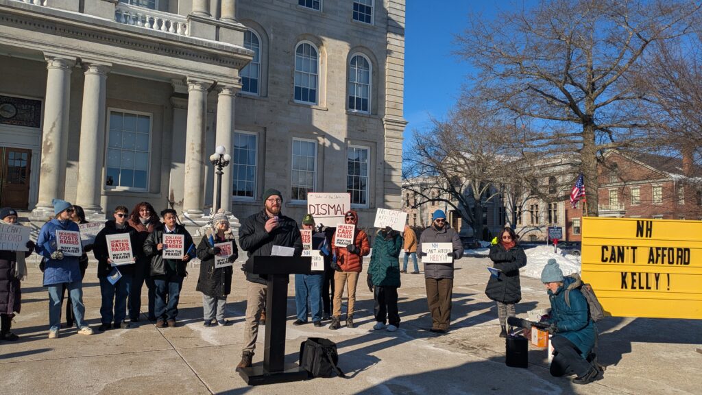 A dozen people holding signs and about the many problems of the state, with one speaking. There's also a larger sign reading "NH Can't Afford Kelly!"